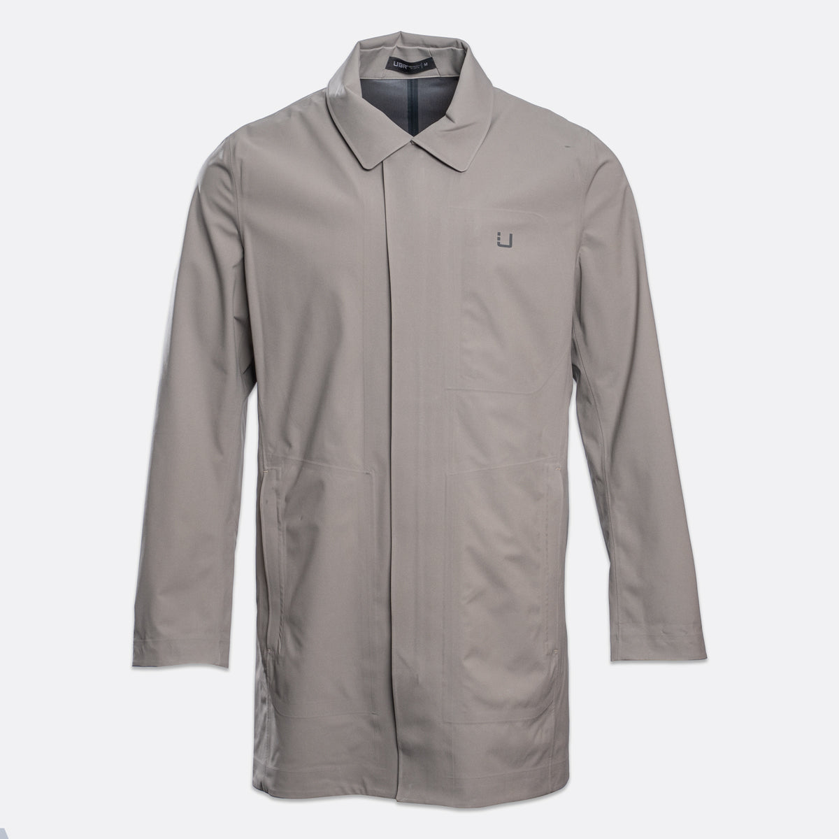 A light gray utility jacket with a pointed collar, long sleeves, and a small square logo patch on the left chest, displayed against a light gray background. UBR Jacket available at Sakks Menswear in Southampton.
