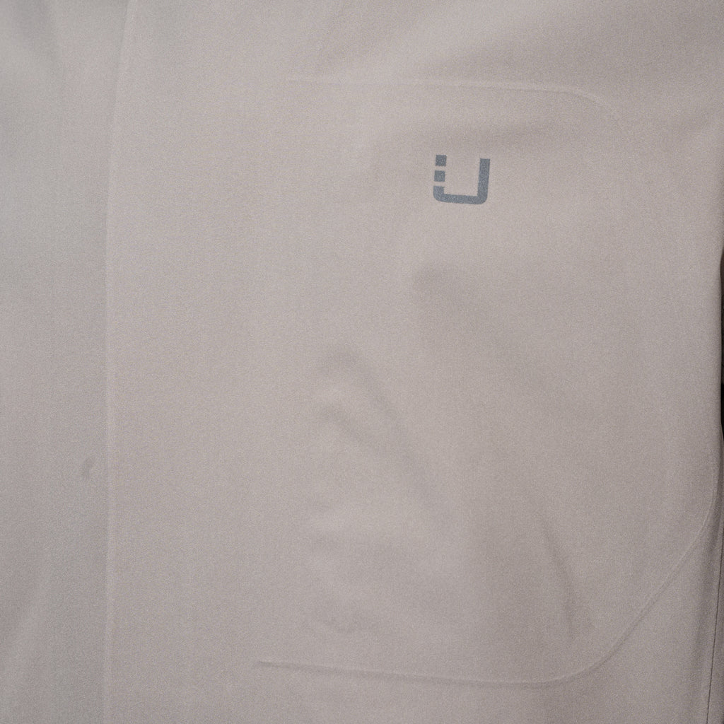 Close-up of a light gray or off-white fabric surface with a small gray stylized 'U' logo near the upper right area. UBR Jacket available at Sakks Menswear in Southampton.