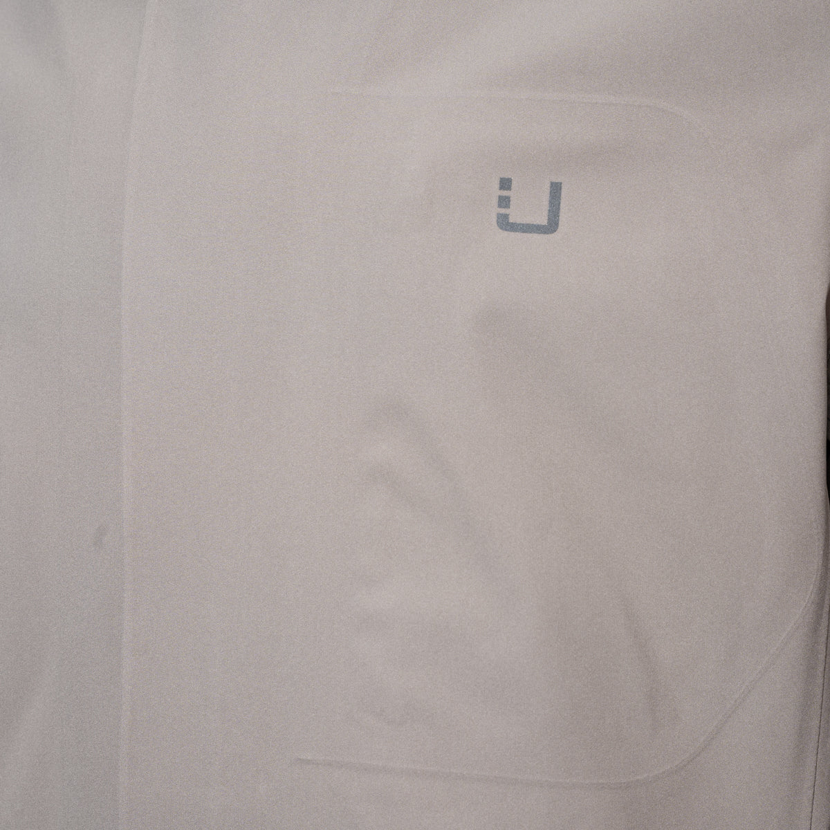 Close-up of a light gray or off-white fabric surface with a small gray stylized 'U' logo near the upper right area. UBR Jacket available at Sakks Menswear in Southampton.
