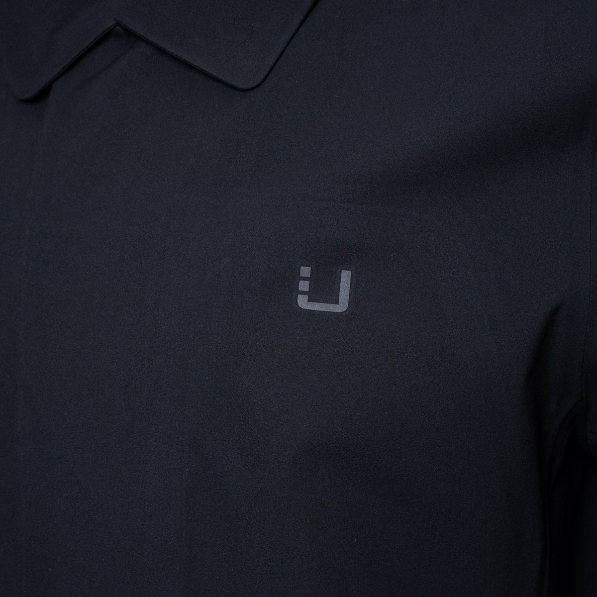 Close-up of a dark navy polo shirt with a small light gray embroidered logo on the chest. UBR Jacket available at Sakks Menswear in Southampton.