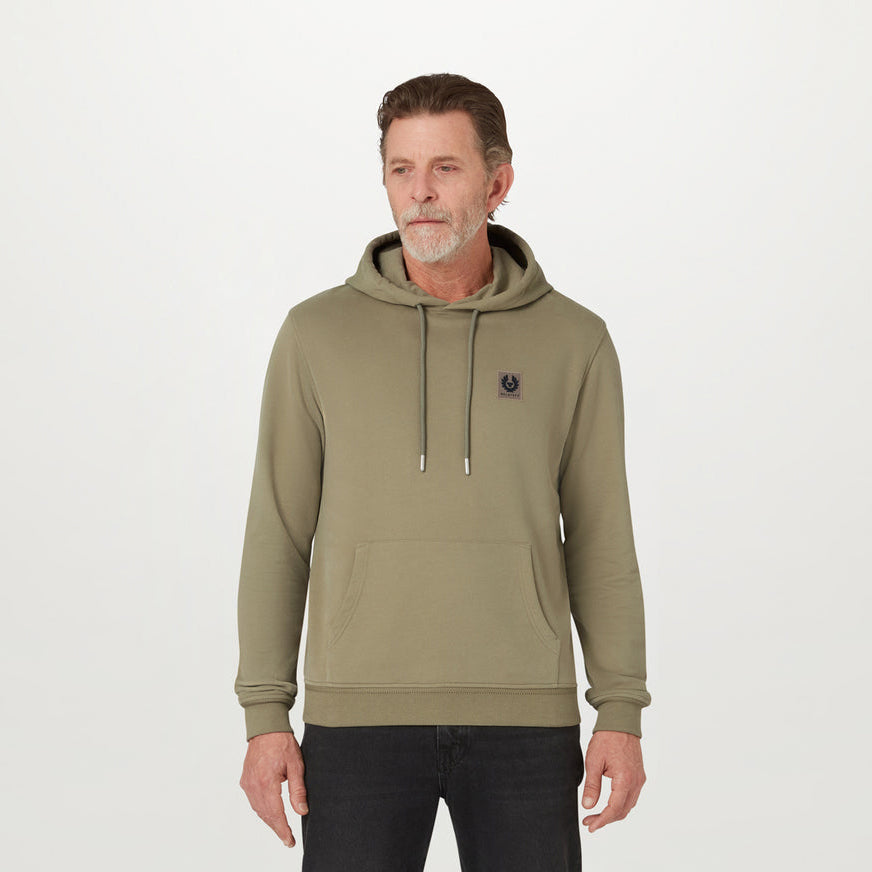 A man wearing an olive green pullover hoodie with a drawstring hood and kangaroo pocket, standing against a plain light gray studio background. Belstaff Hoodie. Sakks Menswear Southampton.