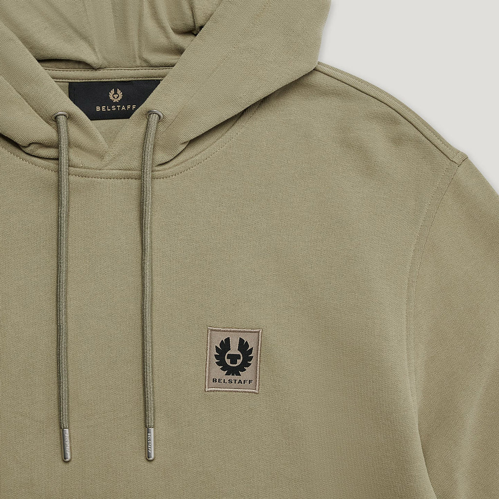 Close-up of a beige hooded sweatshirt with matching drawstrings and a small Belstaff chest patch logo, shown against a light neutral background. Belstaff Hoodie. Sakks Menswear Southampton