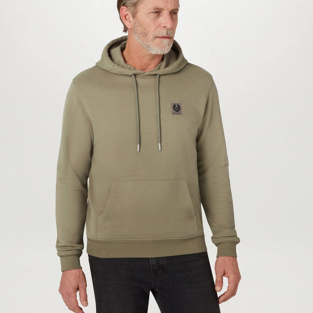 A man wearing an olive green pullover hoodie with a small embroidered logo on the left chest, paired with black jeans, photographed against a light gray studio background. Belstaff Hoodie. Sakks Menswear Southampton.