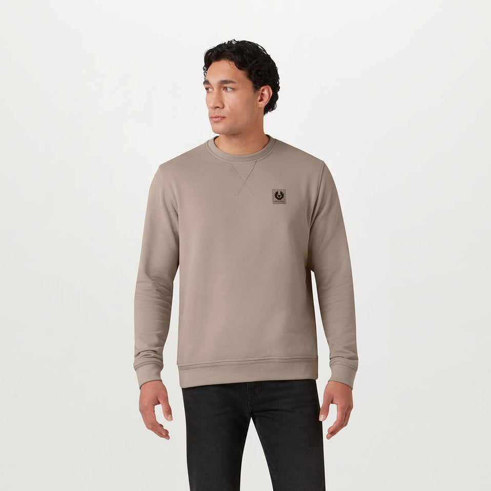 A man wearing a taupe crewneck sweatshirt with a small embroidered logo on the left chest, paired with black pants, standing against a plain white studio background. Belstaff Sweatshirt. Sakks Menswear Southampton.
