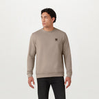 A man wearing a taupe crewneck sweatshirt with a small embroidered logo on the left chest, paired with black pants, standing against a plain white studio background. Belstaff Sweatshirt. Sakks Menswear Southampton.