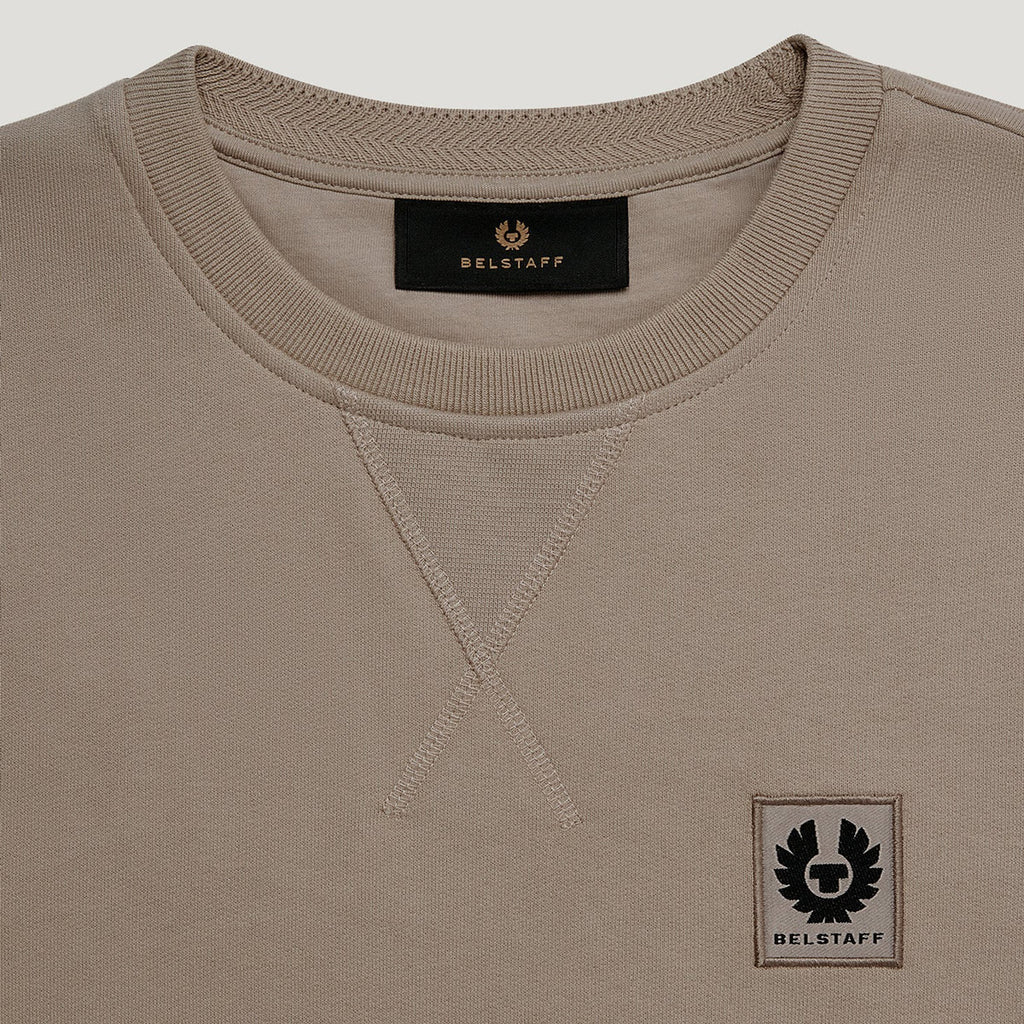 Close-up of a crewneck sweatshirt in taupe beige, featuring a black BELSTAFF brand label inside the collar and a black embroidered winged logo patch on the lower left chest. Belstaff Sweatshirt. Sakks Menswear Southampton.