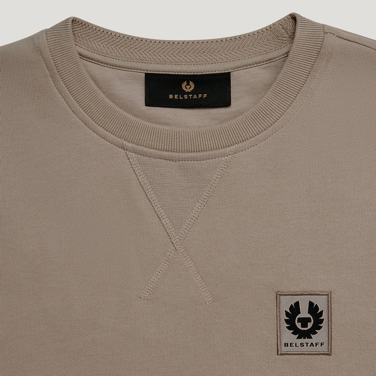 Close-up of a crewneck sweatshirt in taupe beige, featuring a black BELSTAFF brand label inside the collar and a black embroidered winged logo patch on the lower left chest. Belstaff Sweatshirt. Sakks Menswear Southampton.