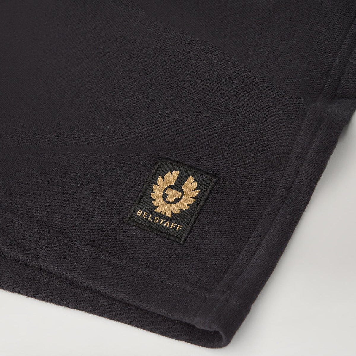 Close-up of a black ribbed fabric corner with a square black patch featuring a gold Belstaff logo and the word BELSTAFF in gold. Belstaff Sweatshorts. Sakks Menswear Southampton.