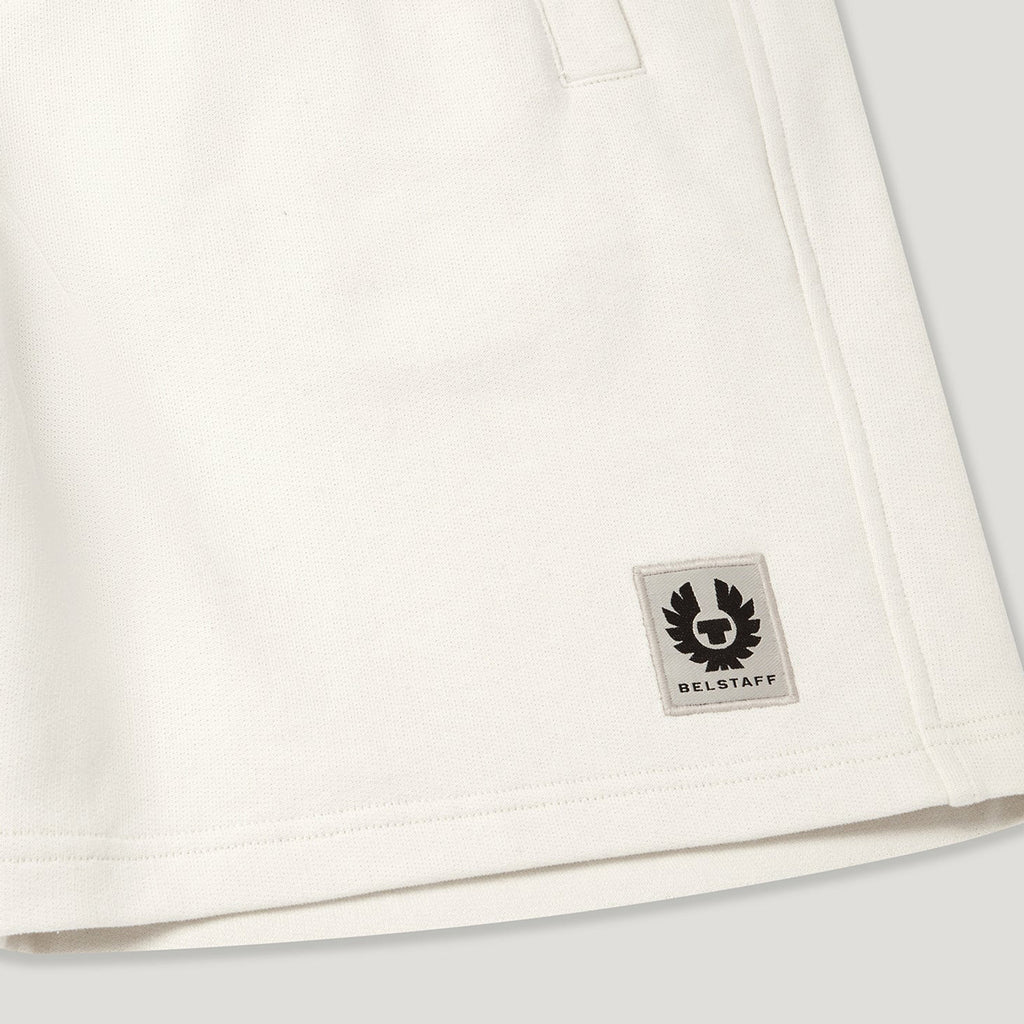 Close-up of an off-white cotton garment, likely a sweatshirt or jacket, showing a small square Belstaff logo patch on the lower right side. Belstaff Sweatshorts. Sakks Menswear Southampton.