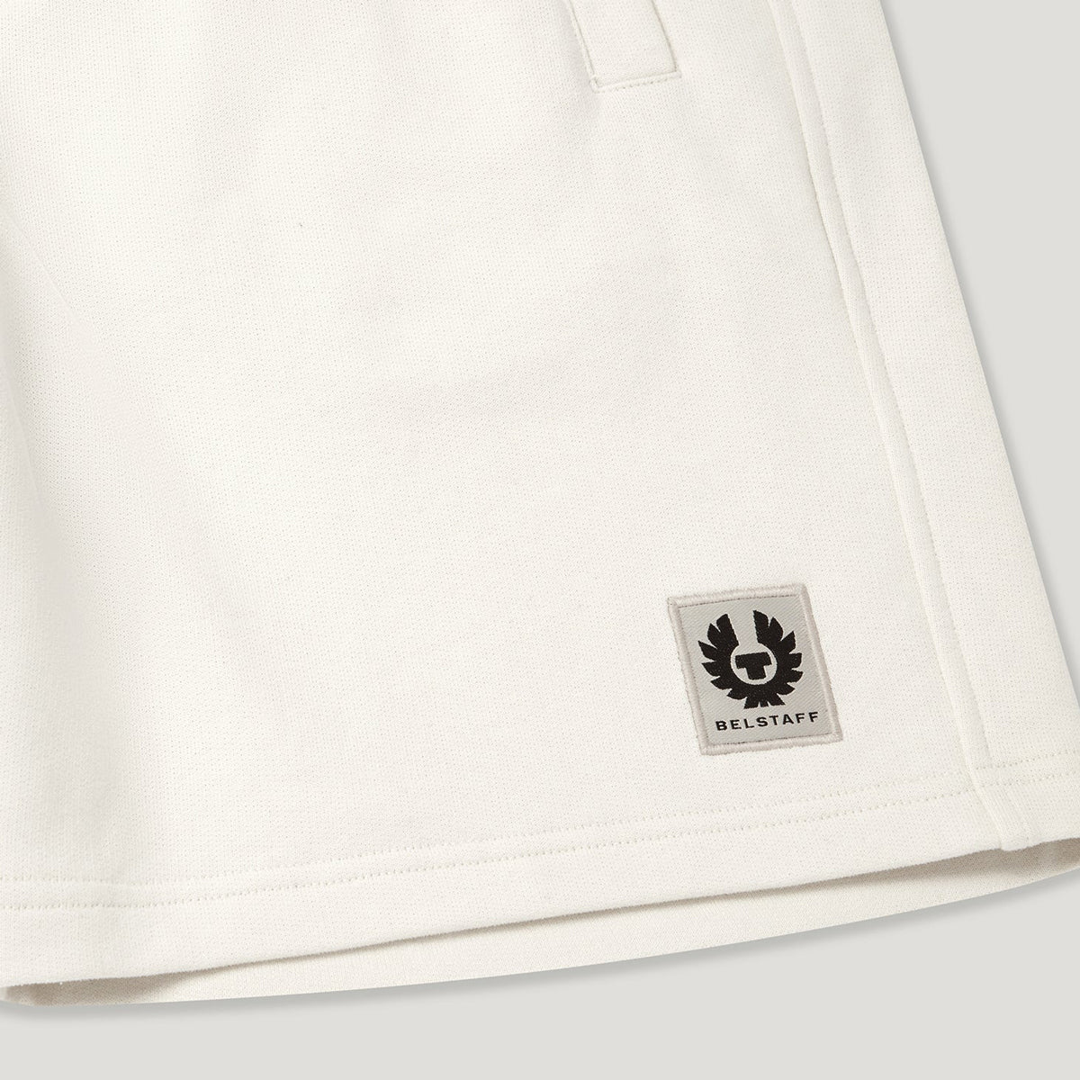 Close-up of an off-white cotton garment, likely a sweatshirt or jacket, showing a small square Belstaff logo patch on the lower right side. Belstaff Sweatshorts. Sakks Menswear Southampton.