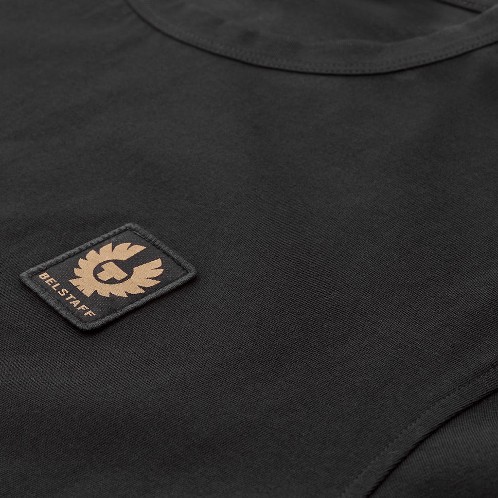 Close-up of a black crew-neck t-shirt featuring a rectangular black patch on the chest with a tan/gold embroidered winged logo and the text 'BEL STAFF'. Belstaff T-Shirt. Sakks Menswear Southampton.