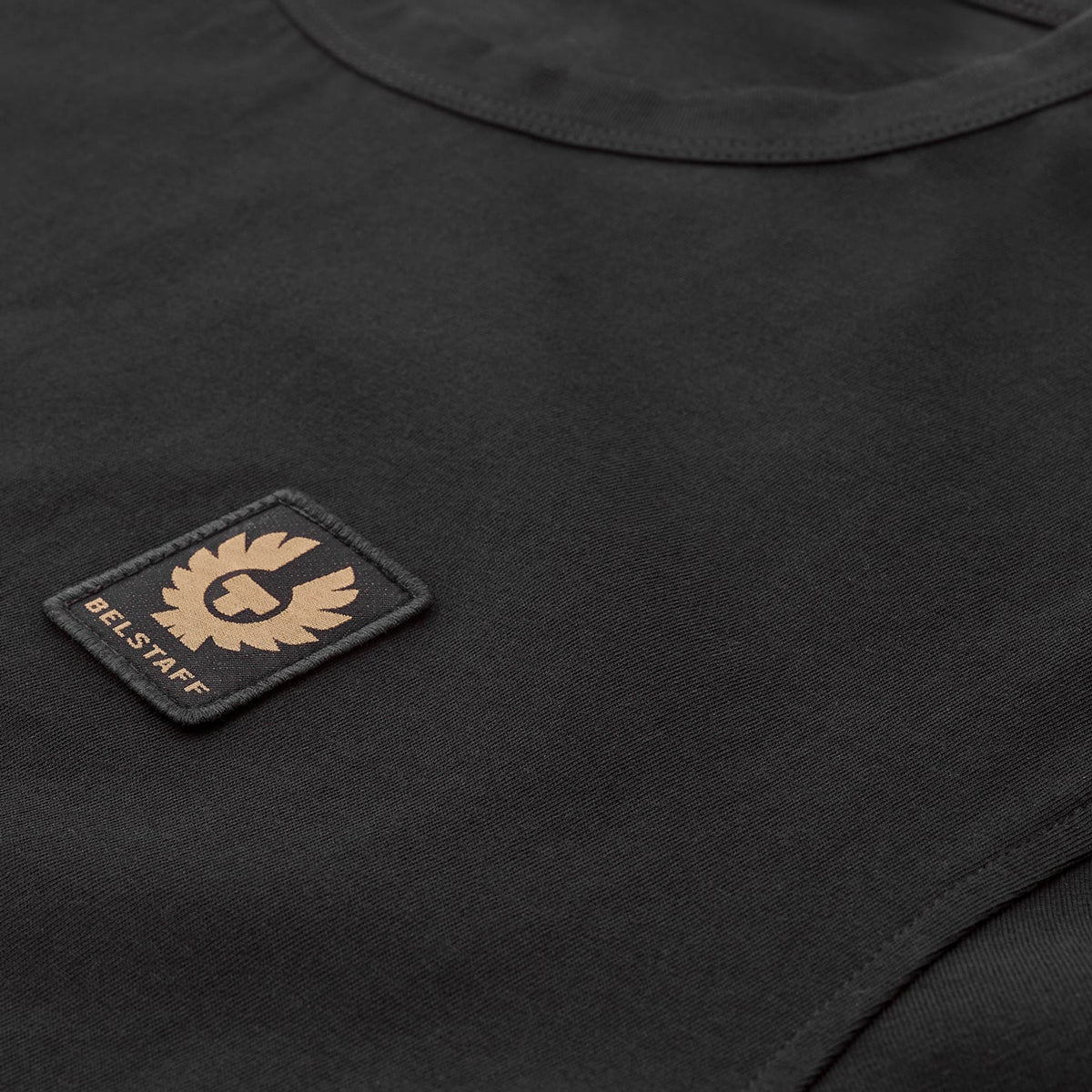 Close-up of a black crew-neck t-shirt featuring a rectangular black patch on the chest with a tan/gold embroidered winged logo and the text 'BEL STAFF'. Belstaff T-Shirt. Sakks Menswear Southampton.