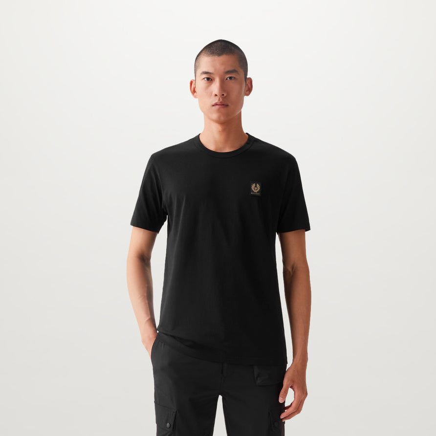 A man wearing a black short-sleeve crewneck t-shirt with a small gold chest logo, paired with black pants, standing against a light gray studio background. Belstaff T-Shirt. Sakks Menswear Southampton.