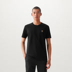 A man wearing a black short-sleeve crewneck t-shirt with a small gold chest logo, paired with black pants, standing against a light gray studio background. Belstaff T-Shirt. Sakks Menswear Southampton.