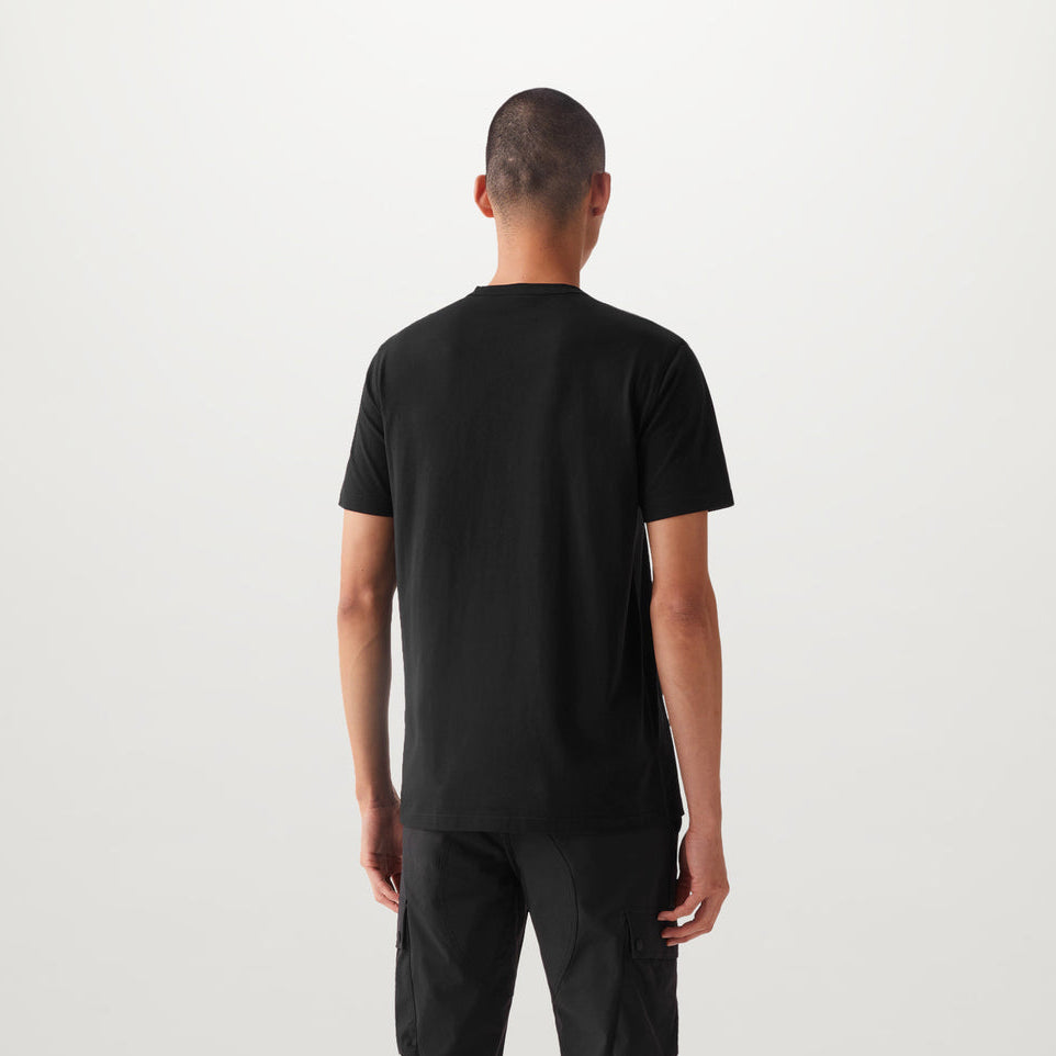 A man viewed from behind wearing a plain black short-sleeve t-shirt and black pants, standing against a light gray studio background. Belstaff T-Shirt. Sakks Menswear Southampton.