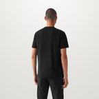 A man viewed from behind wearing a plain black short-sleeve t-shirt and black pants, standing against a light gray studio background. Belstaff T-Shirt. Sakks Menswear Southampton.