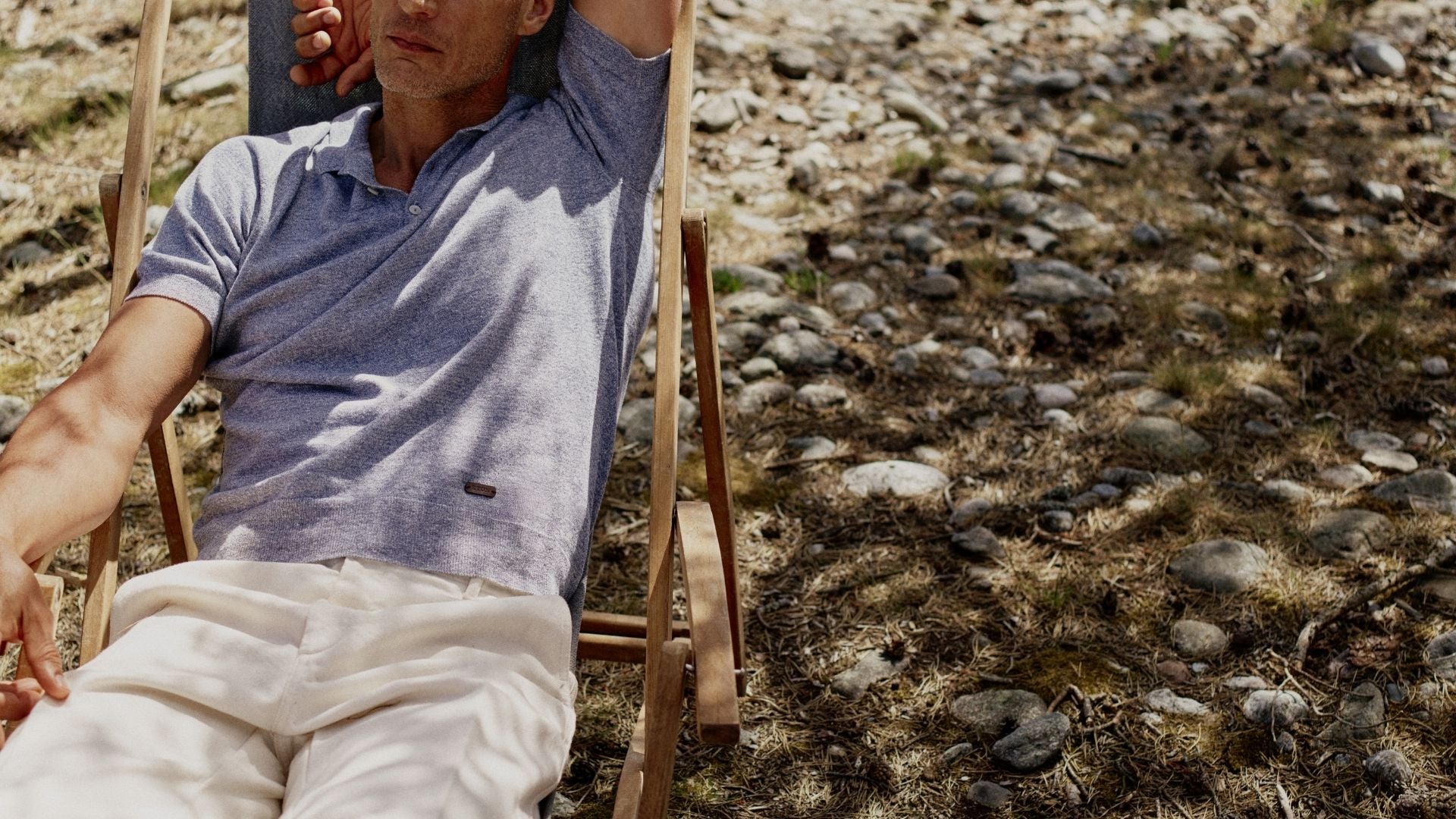 A man reclining in a wooden deck chair outdoors on a rocky, grassy ground, wearing a light blue short-sleeve polo shirt and off-white trousers. Sakks Menswear Designer clothing available in Southampton.